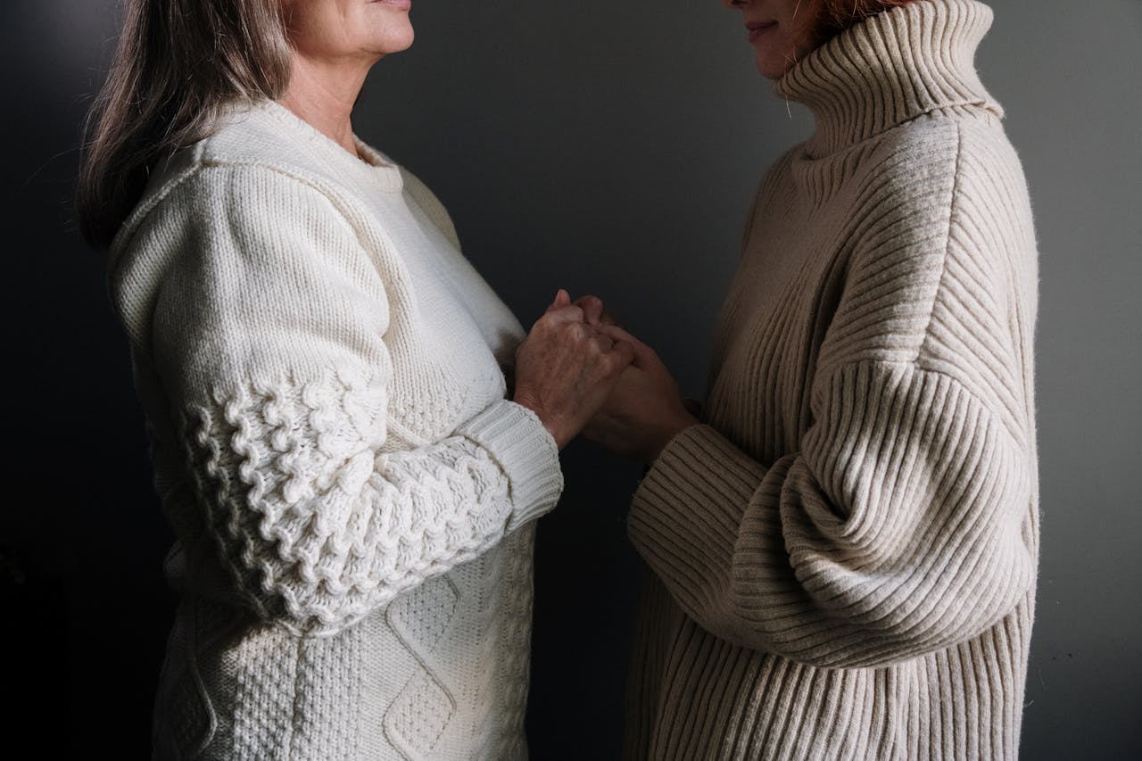 Two women warmly holding hands, showcasing textured knit sweaters indoors.